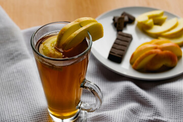 Apple tea in a transparent glass with a handle, the drink is decorated with slices of fresh apple with a yellow peel, next to the plate of sliced fruit and chocolate, everything is on the table on a w