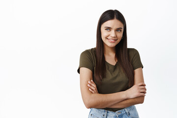 Obraz premium Beautiful young healthy woman smiling, looking away at logo copy space, standing with arms crossed on chest against white background, wearing casual t-shirt