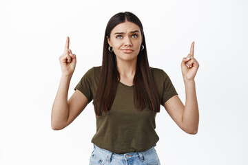 Disappointed young woman with skeptical grimace pointing fingers up, looking at something upsetting...