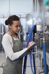 Vertical portrait of young African-American woman holding clipboard while inspecting production...
