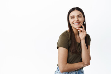 Image of young beautiful female model with long dark hair, looking left and smiling joyfully, staring at logo copy space, touching natural face with light make up, standing over white background