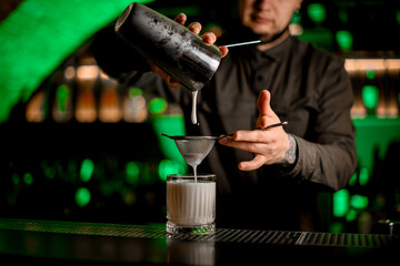 bartender carefully pours cocktail from shaker through sieve into old fashioned glass with ice