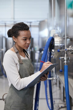 Vertical portrait of young African-American woman holding clipboard while inspecting production quality at industrial food factory, copy space