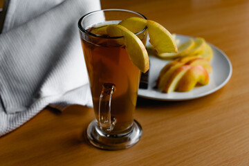 Apple tea in a transparent glass with a handle, the drink is decorated with slices of fresh apple with a yellow peel, next to the plate of sliced fruit and chocolate, everything is on the table on a w