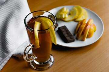 Apple tea in a transparent glass with a handle, the drink is decorated with slices of fresh apple with a yellow peel, next to the plate of sliced fruit and chocolate, everything is on the table on a w