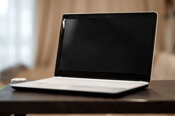 A laptop with a blank black screen in an angular position on a table against a blurred room background. The concept of workplace, office, coworking.