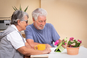 Couple of retired people sitting outdoor on the terrace looking at the same digital tablet. Relaxed moment with coffee cup and book