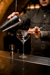 man bartender holds sieve and shaker cup over martini glass