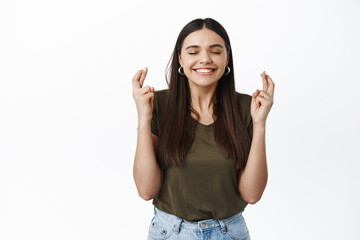 Image of smiling hopeful girl think positive, making wish with closed eyes and big grin, cross fingers for good luck, standing over white background