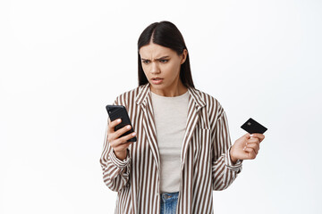 Confused woman staring at her smartphone and holding plastic credit card, cant understand where money on bank account, checking balance or paying online, white background