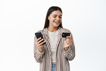 Image of stylish young modern woman smiling, looking at plastic credit card, paying online with mobile phone, shopping in smartphone app, white background