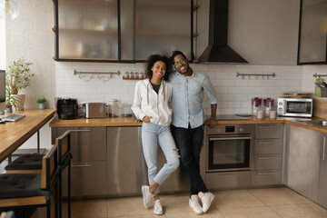 Full length portrait happy African American couple standing in modern kitchen at home, smiling wife...
