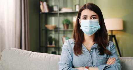 Close up portrait of young beautiful caucasian girl sitting at home on sofa and put on protective mask looking at camera.