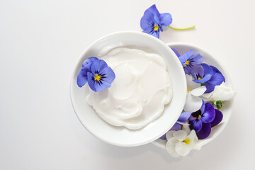 Natural cosmetic ointment and viola or violet flowers in white bowls on a light gray background, copy space, high angle view from above, selected focus