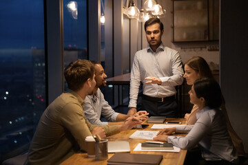 Confident businessman leading late corporate meeting with diverse employees, speaking, explaining...