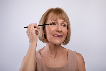 Portrait of lovely senior woman applying eye shadow on her face over light studio background. Beauty and wellness