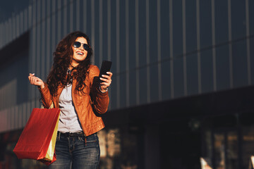 Happy young woman with shopping bags using smartphone on street