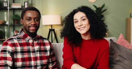Young happy multinational couple sitting at home on sofa turning their head and looking at camera smiling.