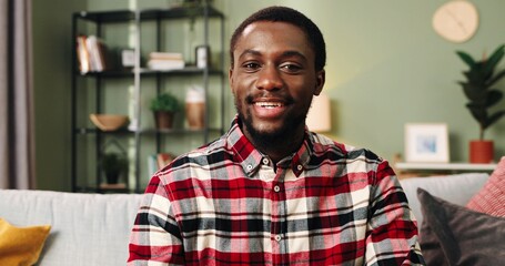 Close up portrait of young happy african american man sitting at home on sofa taking off protective mask and looking at camera smiling.