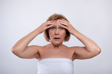 Portrait of terrified senior lady touching her forehead, scared to see new wrinkles on light studio background