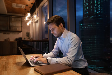 Close up focused businessman using laptop in office at night alone, looking at screen, typing,...