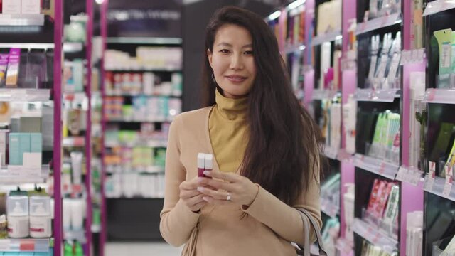 Slow-motion medium portrait of attractive elegant asian woman standing in cosmetics store smiling at camera holding two pink color lipsticks