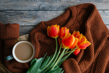 Spring morning coffee. Cozy composition with brown sweater, orange tulips and hot cup of coffee with milk on wooden background. Sunday relaxing, vintage mood and lifestyle concept. Top view copy space