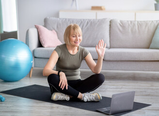 Happy mature woman in sportswear sitting on yoga mat in front of laptop, greeting her personal...