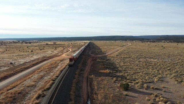 Very long train with containers double stacked on the wagons moving through the dessert over a railway track with long shadows on a partly cloudy summer day. Drone dolly shot