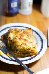 home made lasagna in a white plate and on the wooden desk. blurry background.