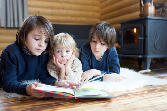 Three sweet children, siblings lying on the floor in little fancy wooden cottage, reading a book, drinking tea