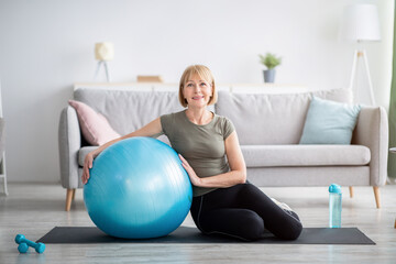 Full length portrait of happy mature woman resting on yoga mat with fitness ball, smiling at camera indoors, free space