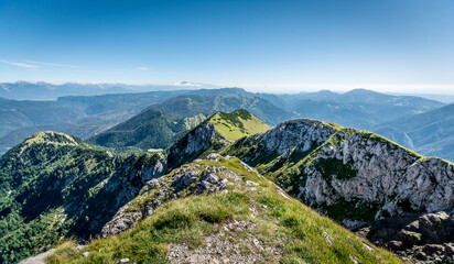 Naklejka na ściany i meble A panoramic view on the Alps from the top of Mittagskogel in Austria. Clear and sunny day. Sharp peaks around. A bit of haze in the valley. Outdoor activity. Lush green slopes with barren peaks. Calm