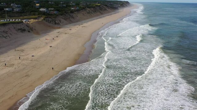 Aerial tilt down shot of people at beach by coastal town against sky, drone flying forward over waves splashing on shore - Cape Cod, Massachusetts