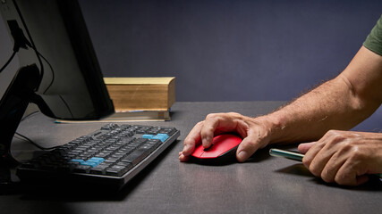 man's hand using the mouse on his computer, in the background books and in his other hand a cell phone