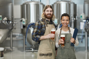 Waist up portrait of two smiling young workers holding beer glasses and looking at camera while...