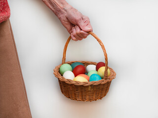 Old, elderly woman holds in her hand a multicolored, painted boiled chicken eggs in a wicker wooden, rustic basket. Traditional elements of a happy Orthodox or Catholic Easter.