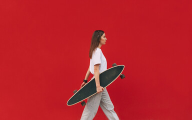 Confident young woman skater in casual outfit is walking in front of an isolated red wall with a copy space and holding her skateboard.