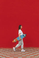 Vertical full-length photo of a fashionable female skater walking the street in front of a red wall, holding her skateboard. © bodnarphoto