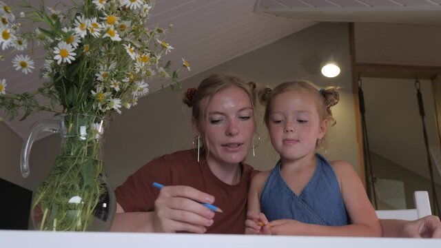 Mom and her daughter are drawing together with colored pencils at home. Mother of the blonde teaches her cute daughter to draw while sitting at the table. Concept: mother's day. 