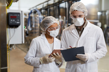 Waist up portrait of two workers wearing masks and lab coats while discussing production at...