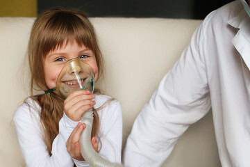 A little girl does inhalation with a spray at home, a doctor is nearby. Inhaler for children from...