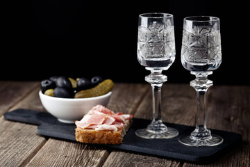vodka in glasses on a black shale snack tray black bread with bacon pickled olive cucumbers on a wooden table
