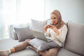 Online Credit Concept. Cheerful Muslim Girl In Hijab Celebrating Success With Card And Laptop At Home, Sitting On Sofa, Raising Fists In Excitement. Arab woman making online purchase on laptop.