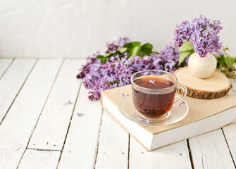 Romantic background with a cup of tea, lilac flowers and a book over a white wooden table. Leisure concept, spring breakfast