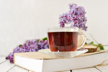 Romantic background with a cup of tea, lilac flowers and a book over a white wooden table. Leisure concept, spring breakfast