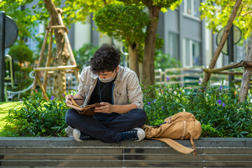 Young asian man with protective mask is sitting outside writing on leather notebook during an epidemic.