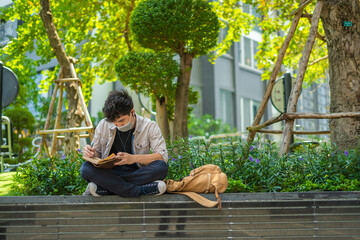 Young asian man with protective mask is sitting outside writing on leather notebook during an epidemic.