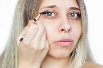 A young beautiful blonde woman using a contour brush while drawing arrows on the eyelids isolated on a white background. A girl makes a makeup herself at home