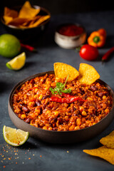 Mexican hot chili con carne in a bowl with tortilla chips on dark background